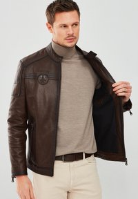 Brown leather jacket with a slim fit, zip closure, stand-up collar, and textured finish. Features a round logo on the chest and side pockets.