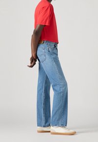 Röd bomulls-t-shirt i kombination med ljusblå raka denimjeans och vita sneakers med beige sulor, visad från sidan.