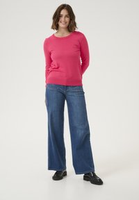 Pull en tricot rose avec un col rond, associé à un jean en denim bleu à jambes larges et des mocassins noirs, sur un fond neutre.