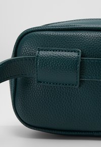 Coin de sac en cuir texturé vert foncé avec un détail de boucle de sangle cousue sur un fond uni clair.
