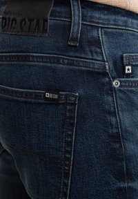 Jeans de mezclilla azul oscuro con un acabado texturizado, costuras prominentes y una etiqueta de cuero negro en la cintura. Dos bolsillos traseros.