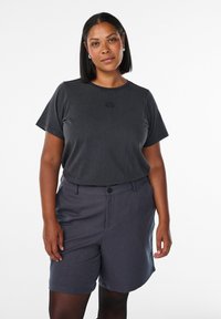 Kvinna med medium hudfärg och rakt svart hår står klädd i en svart kortärmad skjorta och grå skräddarsydda shorts mot en vit bakgrund.