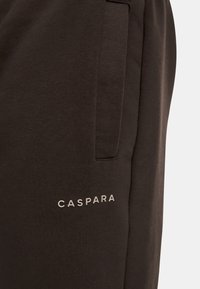 Mörkbruna sweatpants med sidfickor, prydda med en synlig logotyp "CASPARA" i ljusgrått längs nedre vänsterbenet. Mjuk textur.