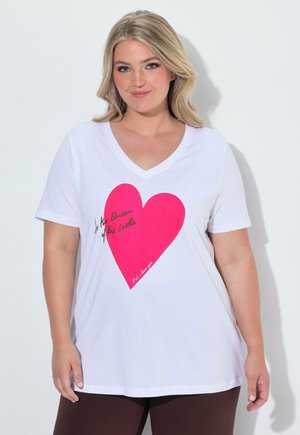 Woman with blonde hair wearing white V-neck T-shirt featuring large pink heart and handwritten text, standing against light background.