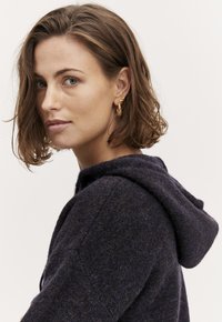 Hooded tmavomodrý pletený sveter s textúrovaným povrchom. Modelka nosí náušnice v tvare zlatej listu. Vlasy sú upravené do voľných vĺn.