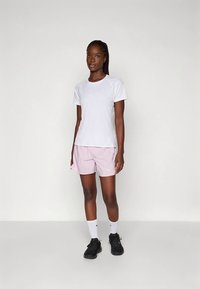 Bílé tričko s krátkým rukávem, růžové sportovní šortky, černé tenisky a bílé ponožky s logem; postoj ve stoje na neutrálním pozadí.