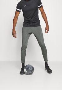 Camiseta deportiva negra con rayas blancas, combinada con pantalones ajustados grises que presentan un logo de Nike; el jugador se encuentra junto a un balón de fútbol en blanco y negro.