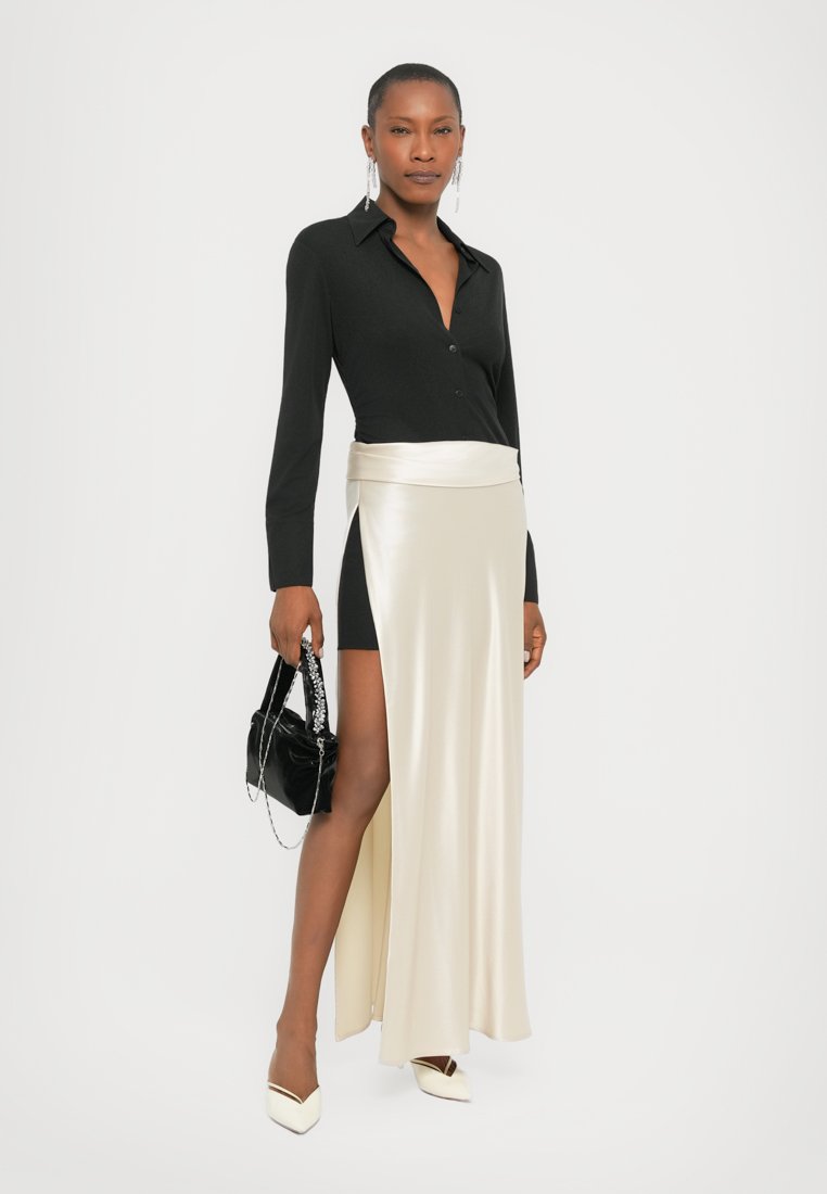 Blouse noire à manches longues, jupe satin crème avec une fente haute, dotée d'une large ceinture. Escarpins blancs à bout pointu et petit sac à main noir.