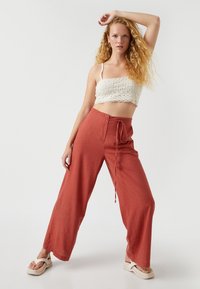 Pantalones ligeros de pierna ancha en color óxido, con una cinturilla ajustable con cordón, confeccionados con una tela texturizada y transpirable; combinados con un top corto de encaje blanco.