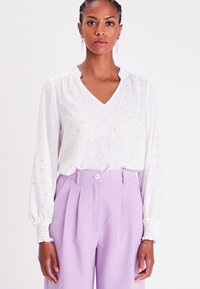 Chemisier blanc avec un motif de pois argentés, décolleté en V, et col froncé, associé à un pantalon lilas taille haute avec des plis et un bouton.