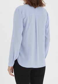 Blouse bleu clair à manches longues avec un ourlet arrondi, des poignets à boutons et un détail plissé dans le dos. Fabriquée en tissu léger et lisse.