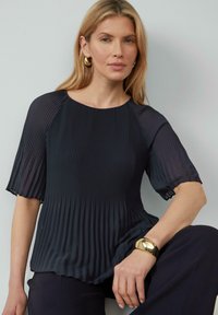 Haut plissé marine en tissu léger, avec des manches courtes et un col rond. Accentué par un bracelet en or au poignet.