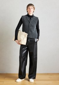 Hallide hall villane vest koos nööpidega musta kõrge kaelusega pluusi peal, millele on sobitatud mustad nahast laiad püksid ja valged tossud. Hoides kreemjat käsitaskut.