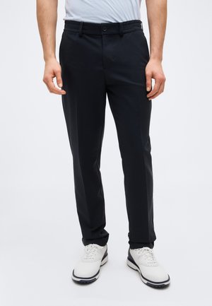 Mann trägt eine schwarze maßgeschneiderte Hose, weiße Golfschuhe und ein hellblaues Hemd und steht vor einem schlichten Hintergrund.