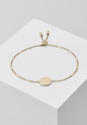 Bracelet chaîne en or délicat avec un centre ovale lisse et un fermoir coulissant ajustable reposant sur un bloc blanc.