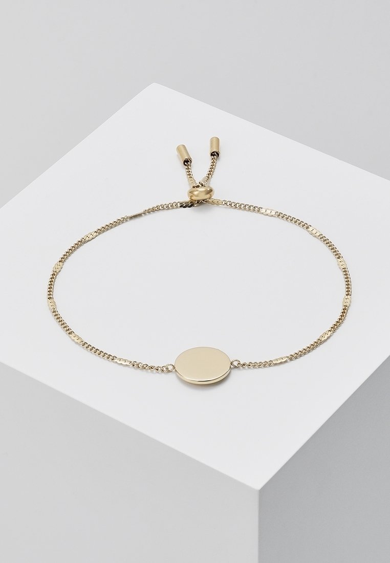 Bracelet chaîne en or délicat avec un centre ovale lisse et un fermoir coulissant ajustable reposant sur un bloc blanc.
