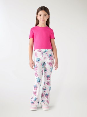 Ragazza giovane che indossa una maglietta rosa acceso a maniche corte con pantaloni bianchi decorati con personaggi dei cartoni animati blu e rosa, in piedi su uno sfondo bianco.