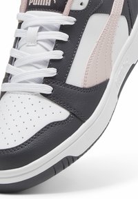 Gros plan sur une chaussure de sport Puma blanche et noire avec une bande rose clair, des lacets blancs, une pointe perforée et des panneaux en cuir texturé.