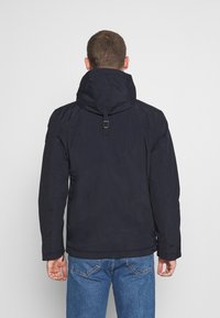Námornická modrá nepromokavá bunda s kapucí, nastavitelným límcem a hladkým povrchem. Nosí se s světle modrými džínami. Má jednoduchý design.