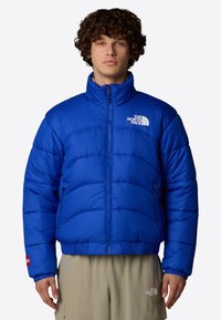 Giacca a vento blu con colletto alto, design trapuntato orizzontale e zip frontale. Logo bianco distintivo sul lato sinistro del petto e dettagli rossi.