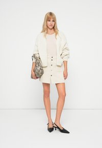 Een romige chunky knit cardigan over een lichtbeige sweater, gecombineerd met een witte button-down rok en zwarte puntige hakken, terwijl ze een gebloemd tasje vasthoudt.