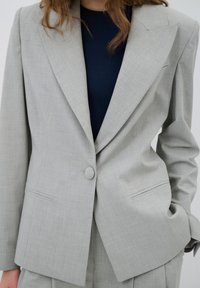 Lichtgrijze op maat gemaakte blazer van een gladde, textuurstof. Kenmerkt zich door een enkele knoopsluiting en notched lapels, gecombineerd met een marineblauwe top eronder.