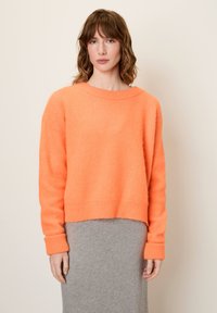 Svetlo oranžni skrajšan pulover z dolgimi rokavi in rebrastimi manšetami, izdelan iz mehke, teksturirane tkanine, v kombinaciji s sivo midi krilom.