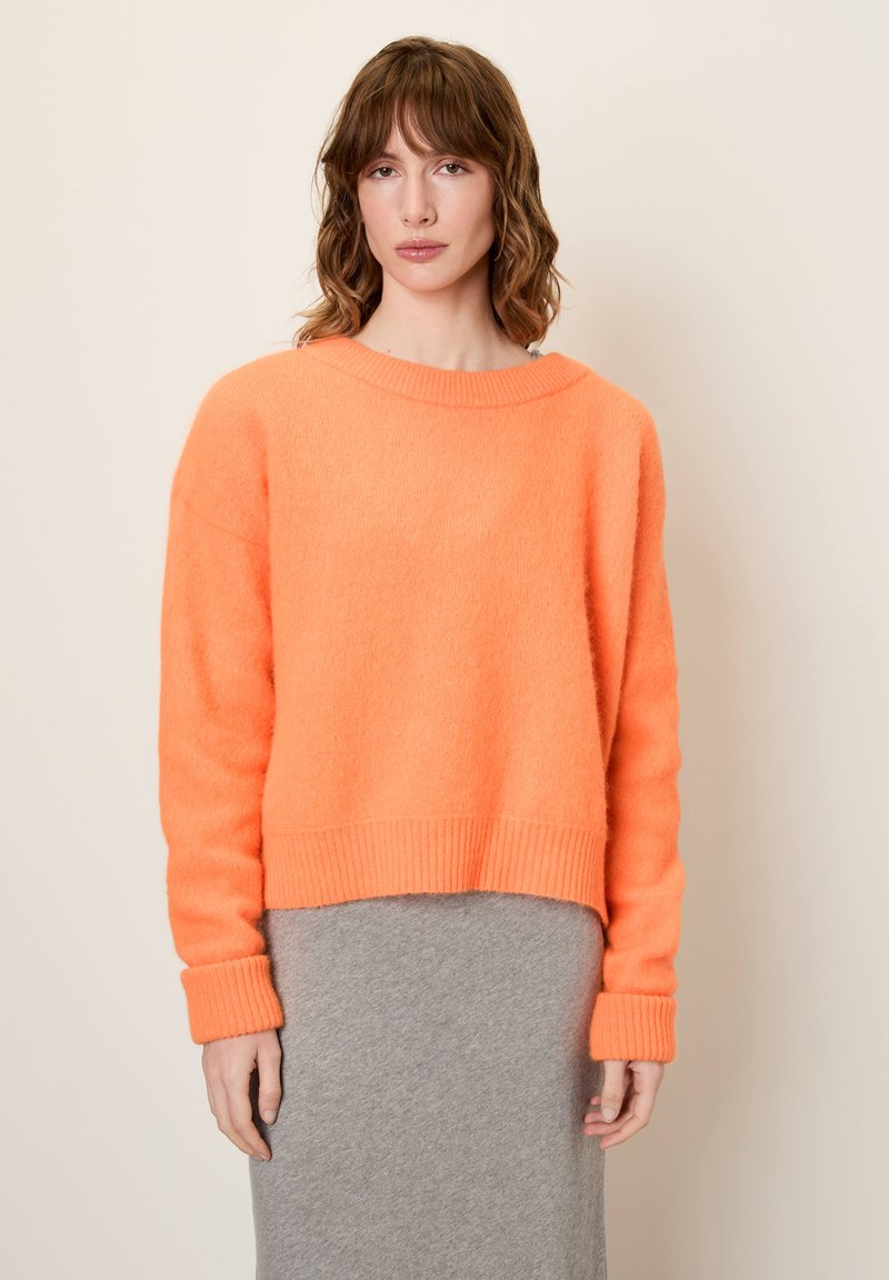 Svetlo oranžni skrajšan pulover z dolgimi rokavi in rebrastimi manšetami, izdelan iz mehke, teksturirane tkanine, v kombinaciji s sivo midi krilom.