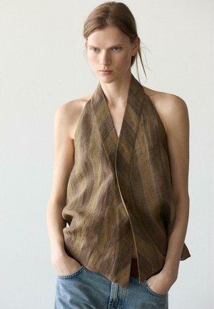 Donna con espressione seria che indossa un top senza maniche a righe marroni con collo all'americana e jeans blu, mani nelle tasche, sfondo neutro.