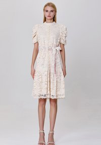 Robe en dentelle crème avec des manches bouffantes, un col haut, une fermeture à boutons, une ceinture à nouer à la taille et un ourlet rufflé. Texture lisse avec des détails de motif floral.