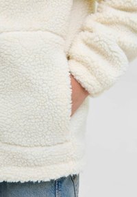 Crème kleurige fleece jas met een gestructureerd oppervlak, voorzien van een zijzak en gathered manchetten. Het materiaal lijkt zacht en comfortabel.