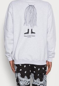 Sudadera gris claro con una ilustración negra de un personaje con cabello largo y botas. Incluye el nombre de la marca, "Makia Clothing Company," debajo.