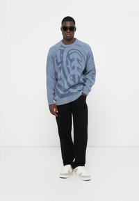Felpa blu a maglia con un motivo strutturato, abbinata a pantaloni neri e sneakers beige. Modello con occhiali da sole scuri.
