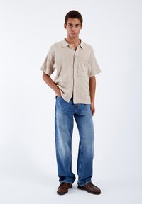 Beige short-sleeve textured shirt with a pocket, paired with light blue loose-fitting jeans and brown shoes. Plain white background.