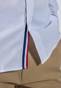 Chemise blanche à boutons partiellement rentrée, montrant des bordures rayées rouge, blanc et bleu sur un pantalon beige.