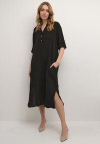 Abito midi in lino nero con scollatura a V, dettagli con bottoni e maniche arrotolate. Presenta tasche laterali e fessure laterali per facilitare i movimenti.