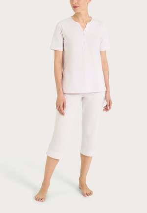 Donna con top rosa chiaro a maniche corte con scollo con bottoni e pantaloni bianchi corti a gamba larga, in piedi a piedi nudi su sfondo semplice.