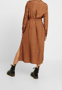 Braunes Midikleid mit Punkten, einem taillierten Bund, langen Ärmeln und seitlichen Schlitzen, kombiniert mit schwarzen Stiefeletten. Glatte Stofftextur.
