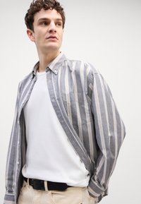 Cămașă cu dungi gri și albă, cu nasturi, având un buzunar pe piept, purtată peste un tricou alb simplu, asociată cu pantaloni deschisi la culoare și o curea neagră.