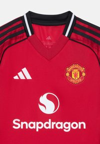 Röd Manchester United-tröja i andningsbart material, med V-hals, svarta ränder på axlarna och broderad logotyp med sponsortext.