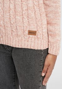 Pull rose clair en maille torsadée avec une étiquette OXMO marron, associé à un jean en denim gris foncé sur le bas du torse et la main d'une personne debout.