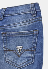 Tasca posteriore di jeans in denim blu con cuciture marroni e una toppa triangolare bianca del marchio Guess.