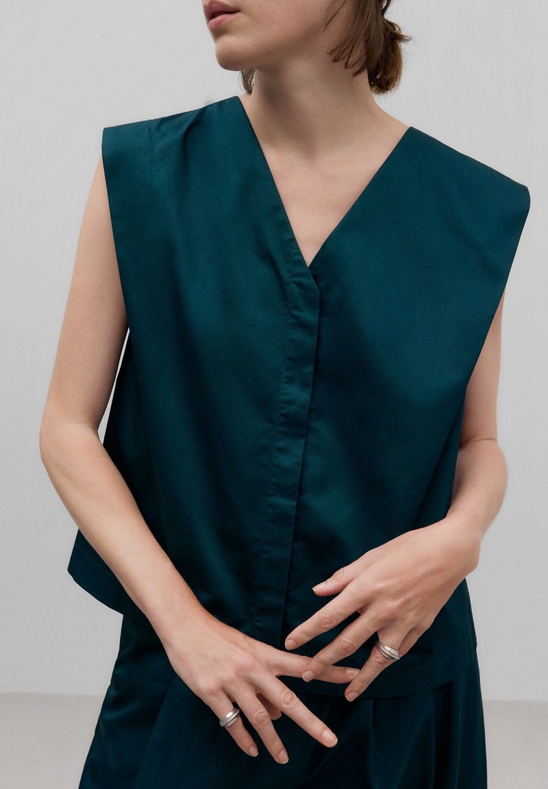 Femme portant un haut sans manches vert sarcelle foncé à col en V et un pantalon assorti, avec des bagues en argent aux doigts et les mains se touchant doucement.
