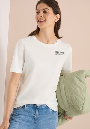 Jeune femme souriante, portant un t-shirt blanc avec le texte « SELFCARE », un jean bleu et tenant une veste matelassée vert clair dans une main.