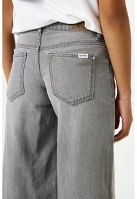 Grijze denim jeans met een losse pasvorm, zichtbare achterzakken en een merklabel aan de taille. De stof heeft een zachte textuur.