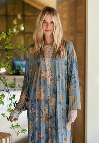 Blonde woman indoors wearing a long floral blue and mustard patterned robe, standing near a green leafy plant.