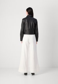 Een persoon met lang haar draagt een zwarte leren jas, witte wijde broek en hakken, staande tegen een eenvoudige grijze achtergrond.