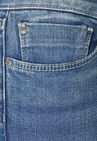 Džínové kalhoty z pohledu zezadu v jasně modré barvě, viditelné švy, malá kapsa s nýty a mírně opotřebovaný vzhled.