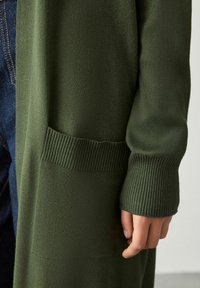 Mørkegrøn cardigan med en blød tekstur, der har ribbet manchet og en frontlomme. Materialet virker let og glat.