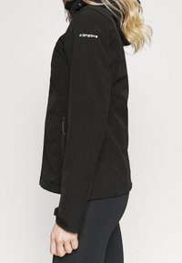 Kvinde i sort softshell-jakke med ICEPEAK-logo på ærmet, lynlås-lomme, justerbar manchet og hætte, vist fra siden mod hvid baggrund.